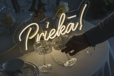 Warm white handwritten LED neon sign "Prieká!" above champagne glasses